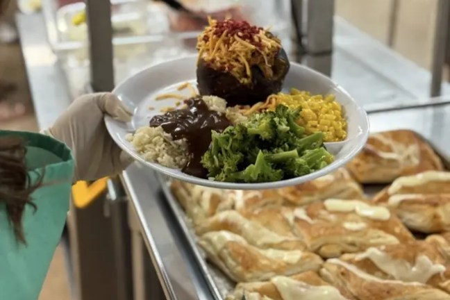 Person holding a plate with steak, broccoli, rice, and corn over trays of pastries.