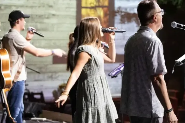 Group of singers performing with microphones on stage, background shows a stained glass window.